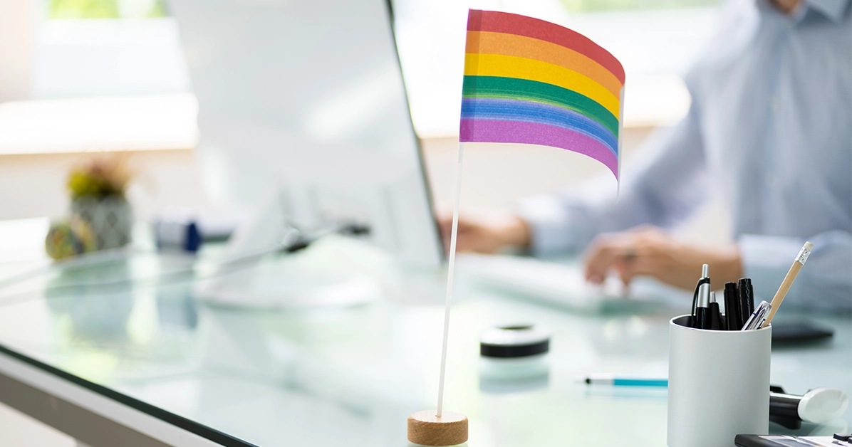 The image shows a rainbow flag on an office desk with a worker using the computer in the background. Blog title: 6 Ways Brands Can Support Allyship That Isn't Rainbow Washing